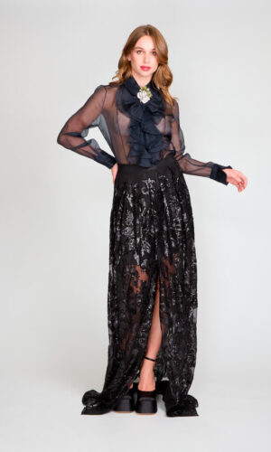 Blusa de gasa negra con vuelos y detalle floral. Falda larga, de encaje de lentejuelas con abertura central.
